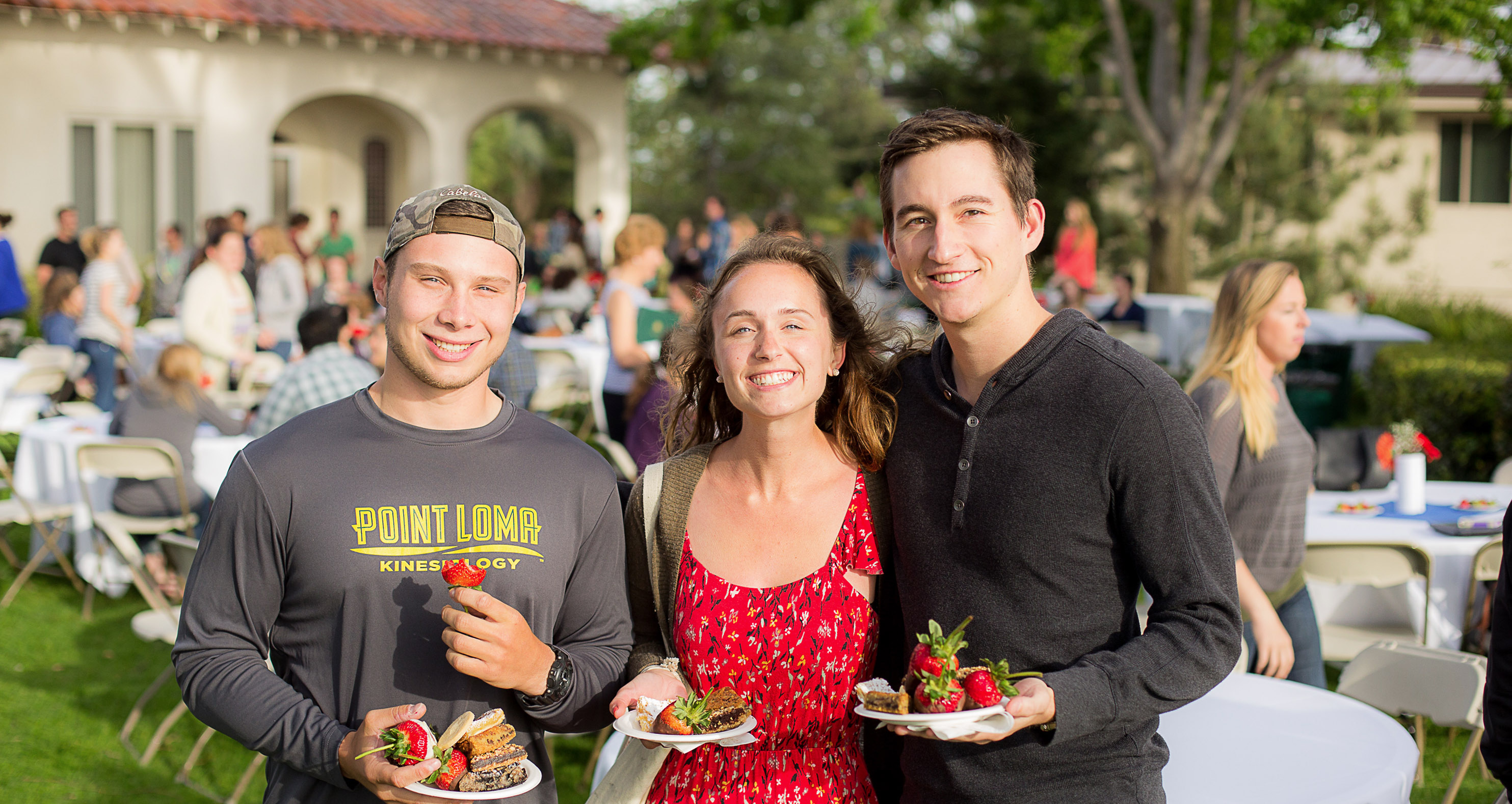 Three friends eat strawberries and dessert at a senior event on alumni lawn