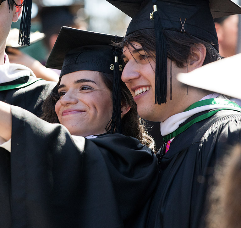Students at PLNU smiling while taking a selfie at graduation in full cap and gown.