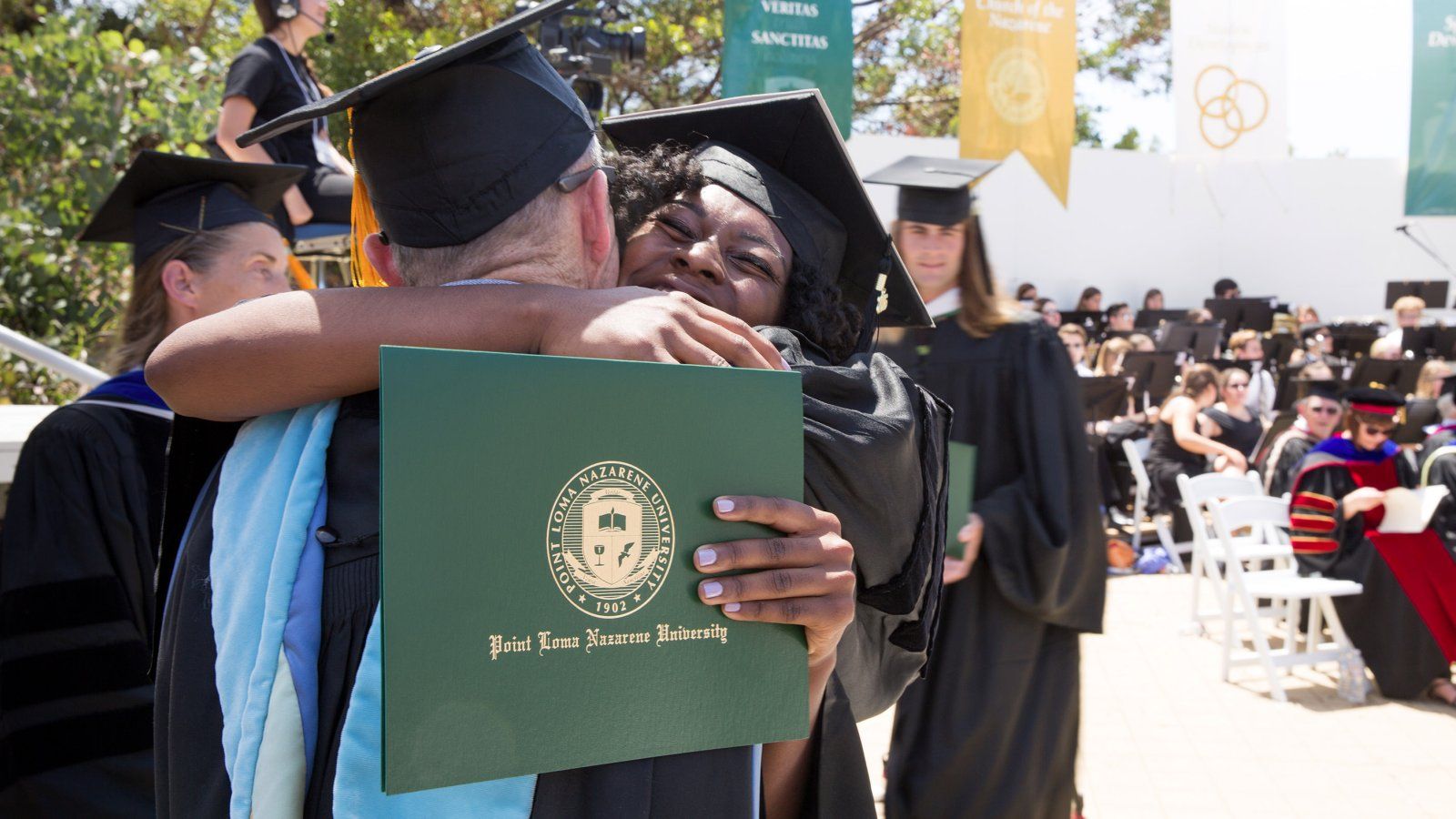 student hugs her professor with diploma in hand, in her cap and gown
