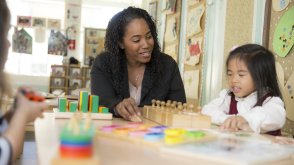 A teacher sits at a table with one of her young students. They are both smiling and are working on a puzzle together.