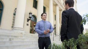 A man in a blue button down is standing in front of San Diego City Hall, talking to a man in a black suit.