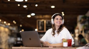 A woman is smiling and sitting in a coffee shop. She is wearing headphones and has an open laptop in front of her.