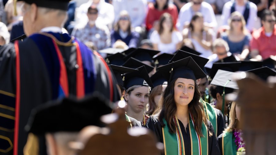 A female college graduate sits at her commencement ceremony wearing a cap and gown. She is smiling while looking at the university's president.