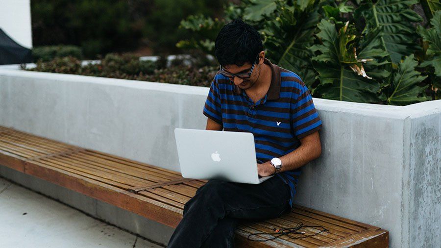 A male student sits on an outside bench and works on his laptop.
