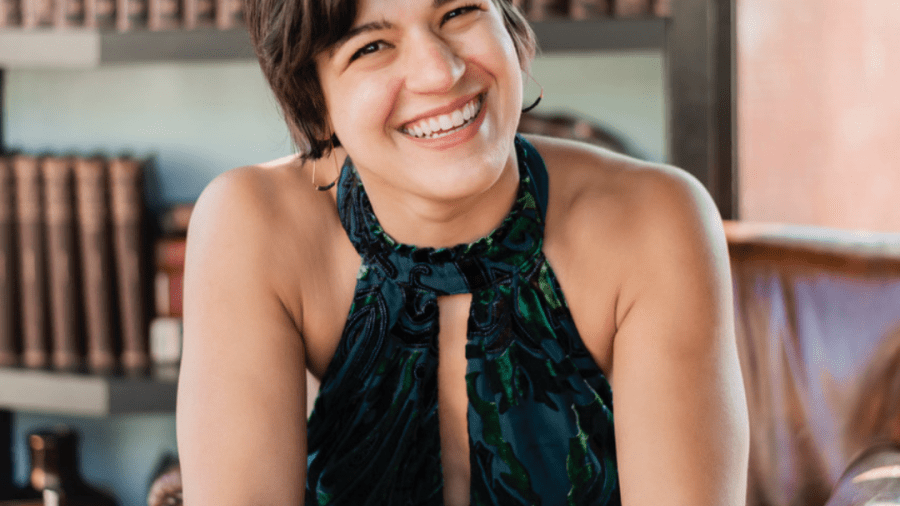 Headshot of Anna Gazmarian seated, smiling at the camera with a blue and green dress on and rows of books behind her.