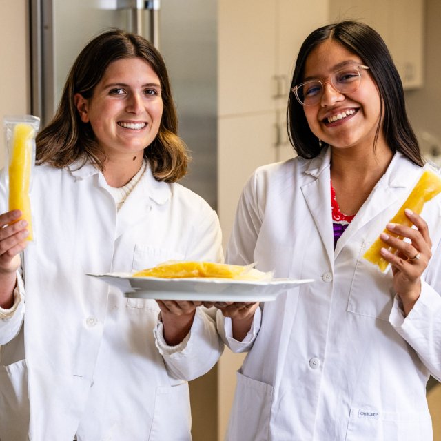 Two PLNU students pose with plate of food in white lab coats set in a kitchen space.