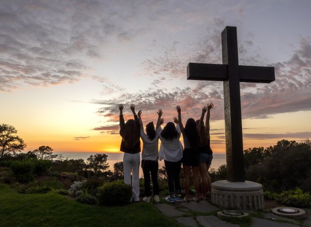 Students raise their hands next to the PLNU cross