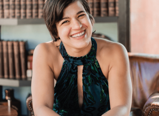 Headshot of Anna Gazmarian seated, smiling at the camera with a blue and green dress on and rows of books behind her.