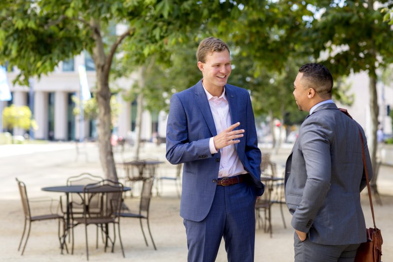 Two business men wearing suits are standing outside on a patio. They are talking and smiling.