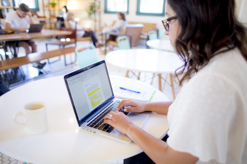 A woman in a white shirt is sitting in a coffee shop. Looking over her shoulder, she is updating her resume on her computer.