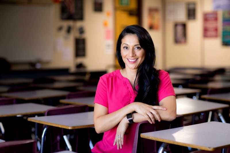 A teacher smiles and sits at a classroom desk