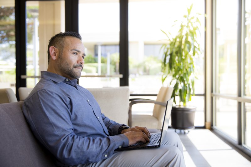 A man in a blue button down and grey pants sits on a couch with a laptop on his lap. He is looking up, thinking whether he should pursue a Master's in Writing or an MFA in Writing.