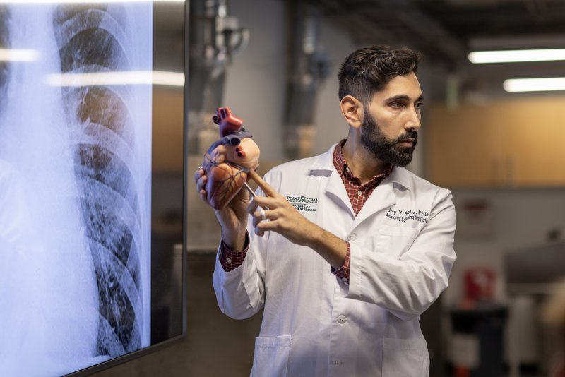 Dr. Joy Y. Balta, wearing a white lab coat, points to a model of a human heart in front of a large medical screen displaying an X-ray image. He appears to be in an educational setting, explaining anatomy concepts to an audience.