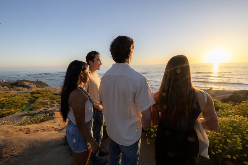 Four students stand on Sunset Cliffs together. They are smiling and looking at the sunset in the horizon.