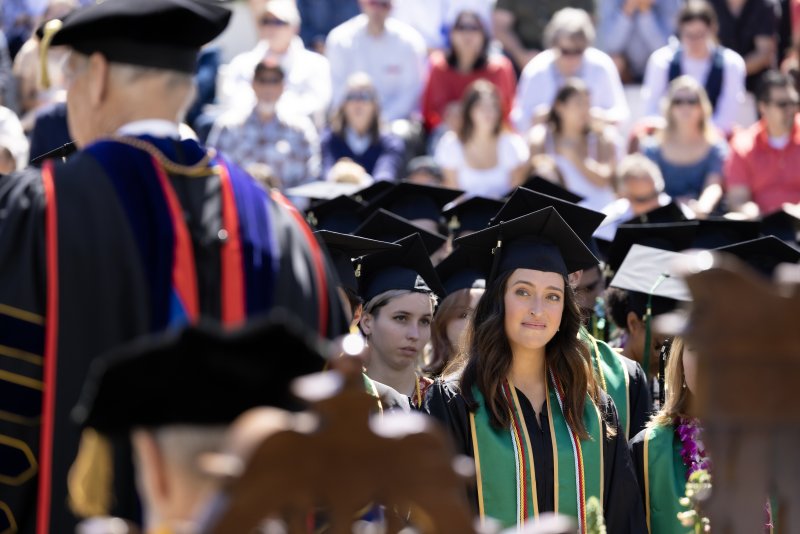 A female college graduate sits at her commencement ceremony wearing a cap and gown. She is smiling while looking at the university's president.