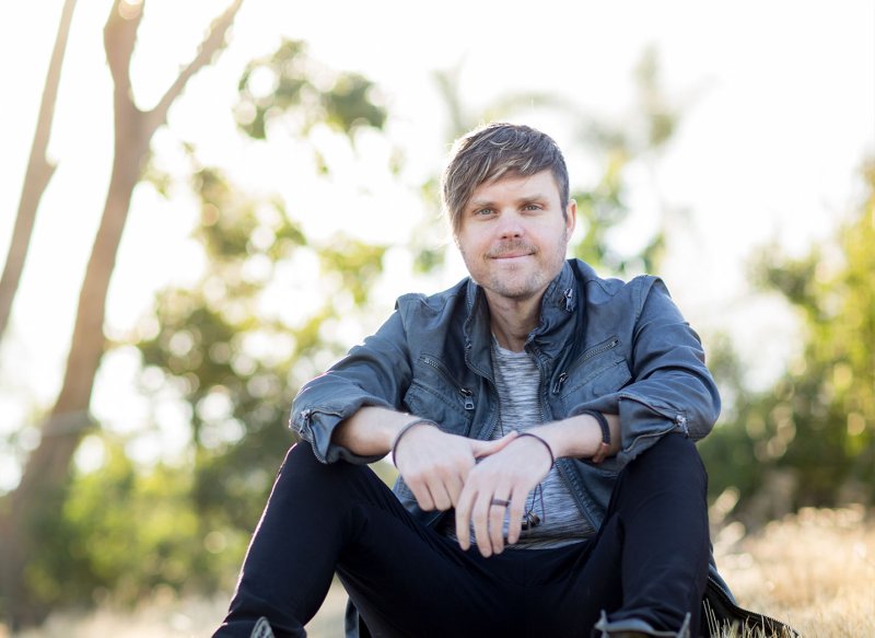 Jason David Sluyter sits in a grass field and confidently smiles at the camera