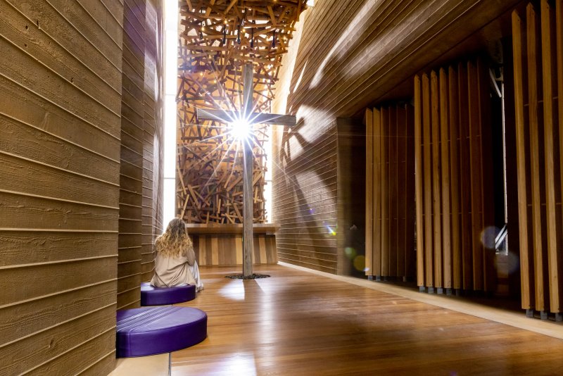A female student sits on a purple cushion in PLNU's Prescott Prayer Chapel. She is looking up at a wooden cross and sunlight is peering through the window behind the cross.