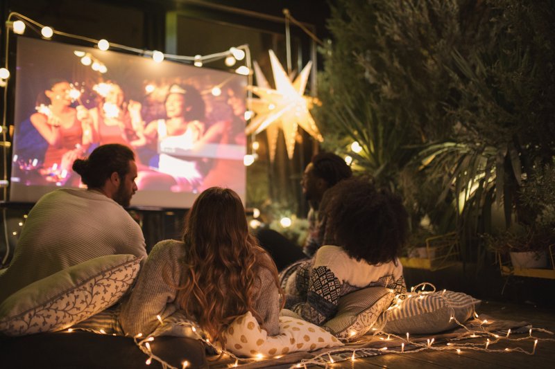 Three people laying on blankets, looking at a movie screen outdoors.